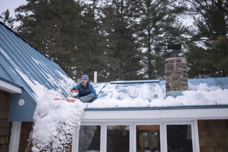 Man shoveling heavy snow off a snow-covered metal roof using a red shovel, with a stone chimney visible in the background