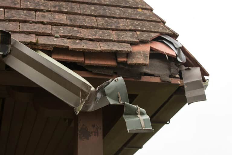 Damaged residential roof corner showing heavily deteriorated shingles, exposed flashing, and a drooping gutter detaching from the soffit