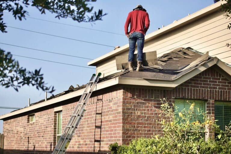 Person standing on the edge of a low-slope residential roof with damaged peeling shingles and an extension ladder propped against the brick wall