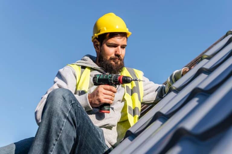Bearded roofer in a yellow hard hat and reflective vest using a cordless drill to repair gray metal roof panels in Harrisburg, PA