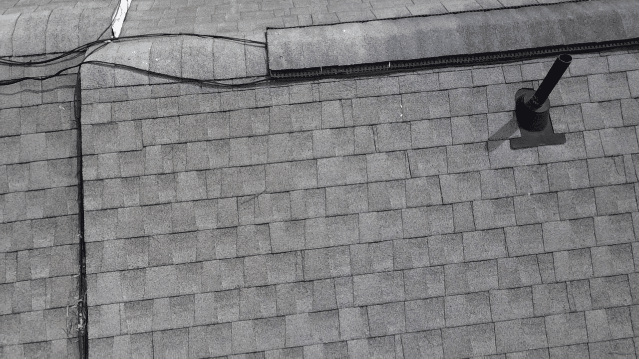 Drone aerial view of a gray asphalt shingle roof showing a ridge vent strip and a black pipe vent