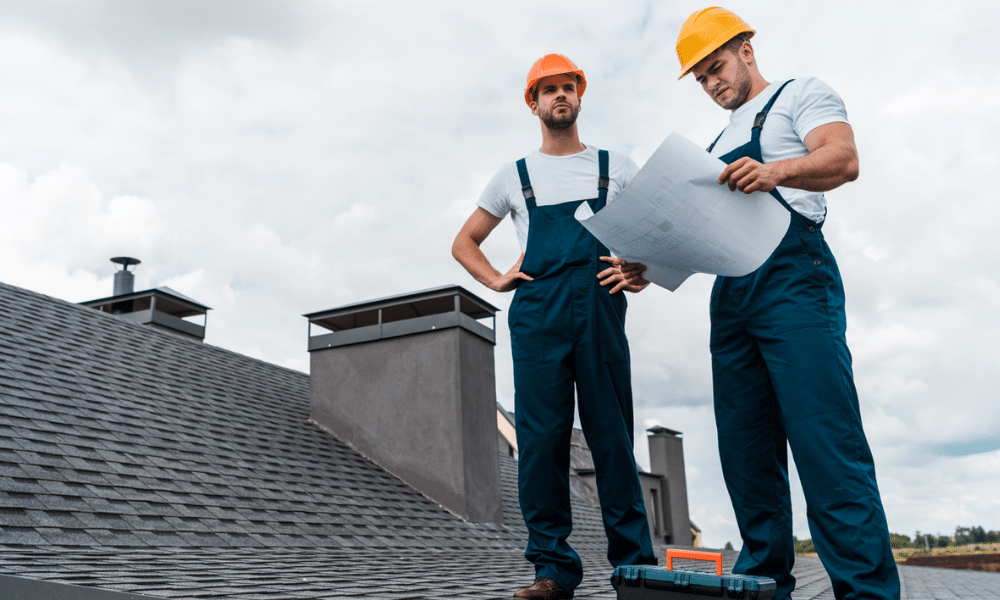 Two roofers in hard hats standing on a gray shingle roof reviewing construction plans together during a roof inspection in Central PA