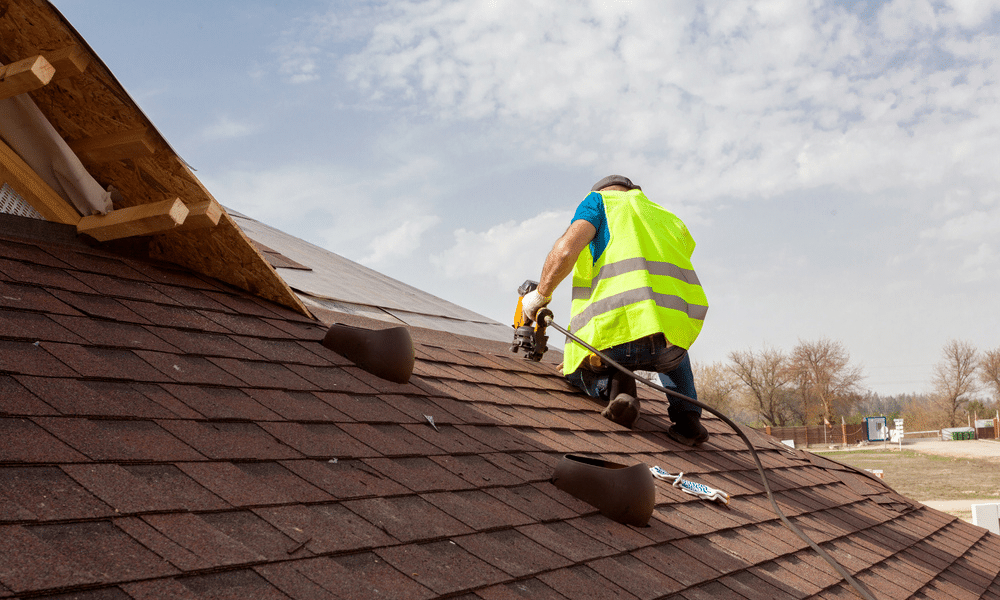 Roofer in a yellow high-vis vest using a pneumatic nail gun to nail dark red shingles near the peak of a home with exposed OSB decking in Central PA