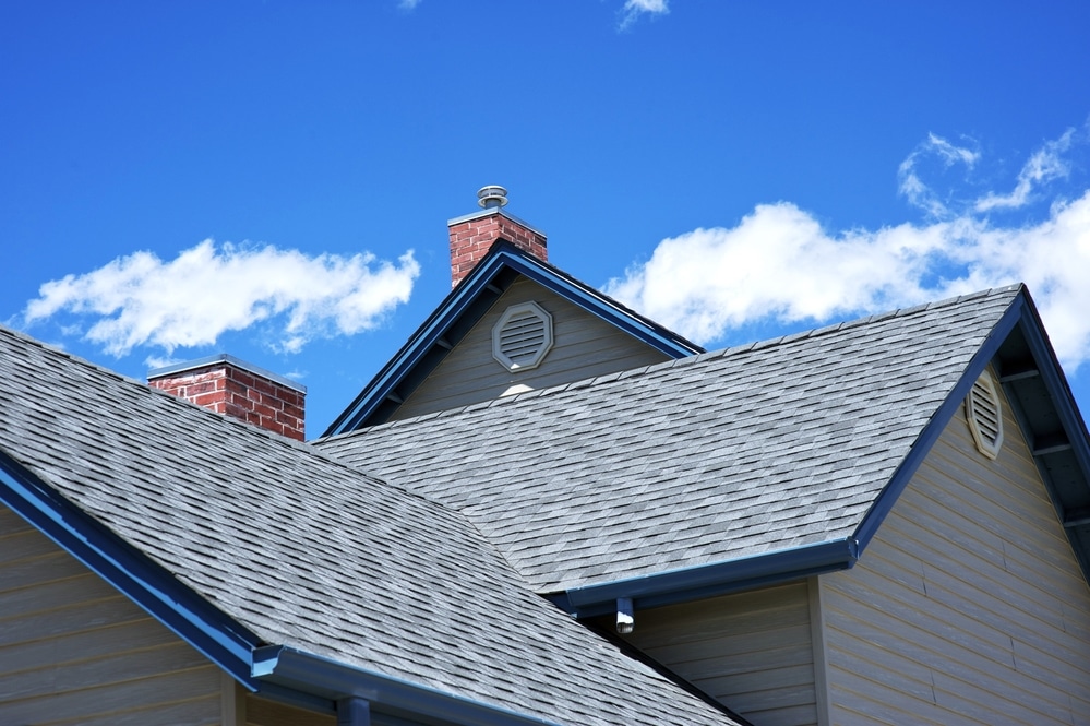 Gray asphalt shingle roof with two gable peaks, a red brick chimney, and circular gable vents against a blue sky